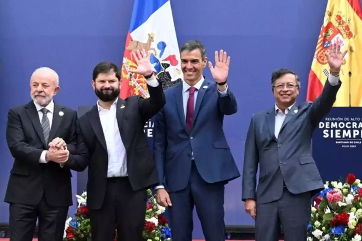 Four men in suits pose smiling and wave with raised hands in front of flags and flowers at an official event.