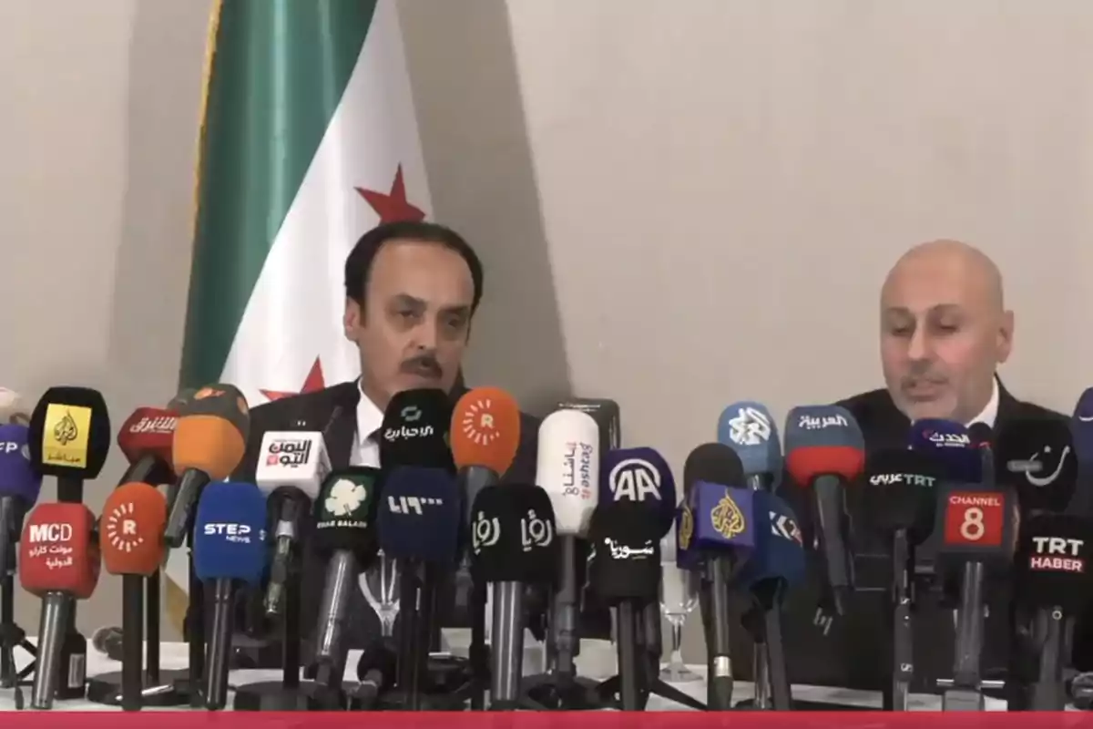 Dos hombres con traje están sentados frente a numerosos micrófonos de diferentes medios de comunicación durante una conferencia de prensa, con una bandera de fondo. Dos hombres con traje están sentados frente a numerosos micrófonos de diferentes medios de comunicación durante una conferencia de prensa, con una bandera de fondo.