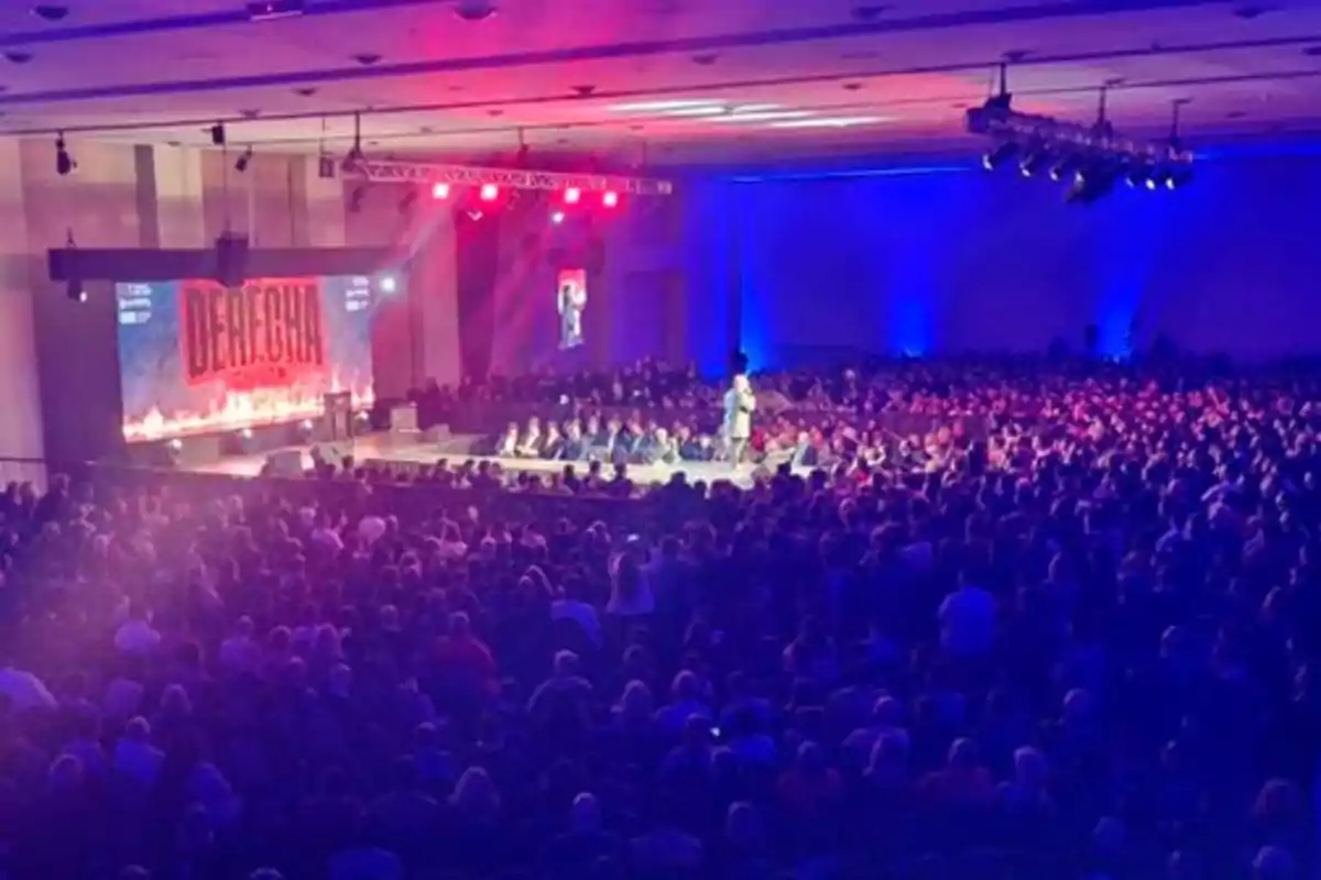 Auditorio lleno de personas asistiendo a una conferencia con luces rojas y azules y un escenario al frente donde una persona está hablando Auditorio lleno de personas asistiendo a una conferencia con luces rojas y azules y un escenario al frente donde una persona está hablando