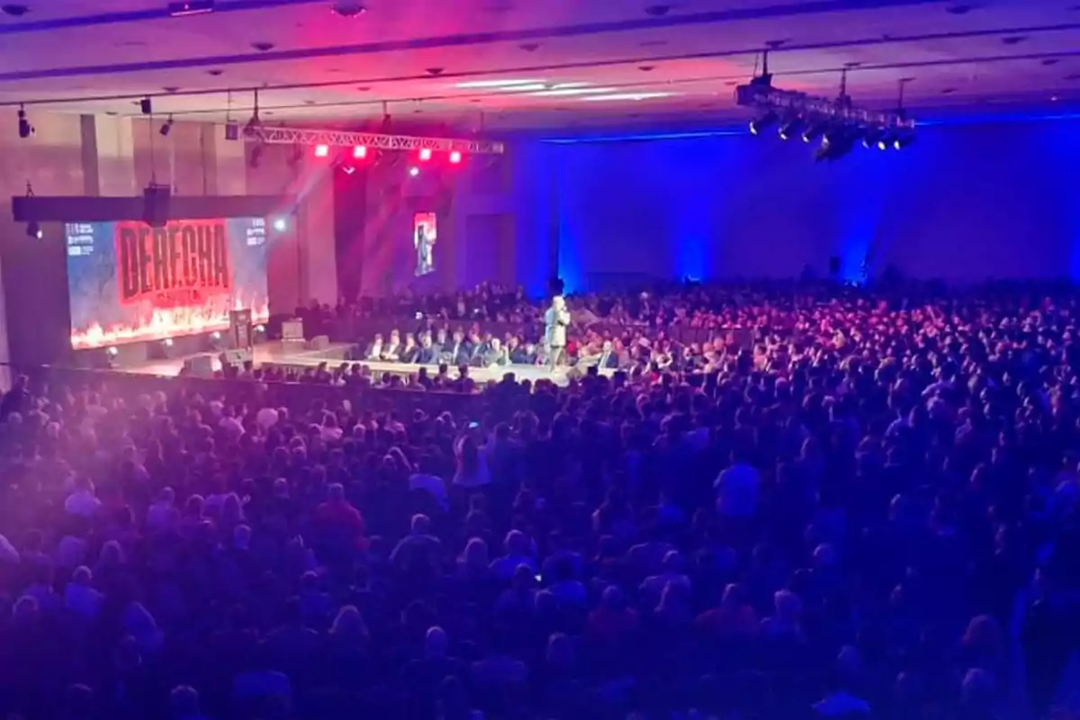 Auditorio lleno de personas asistiendo a una conferencia con luces rojas y azules y un escenario al frente donde una persona está hablando Auditorio lleno de personas asistiendo a una conferencia con luces rojas y azules y un escenario al frente donde una persona está hablando
