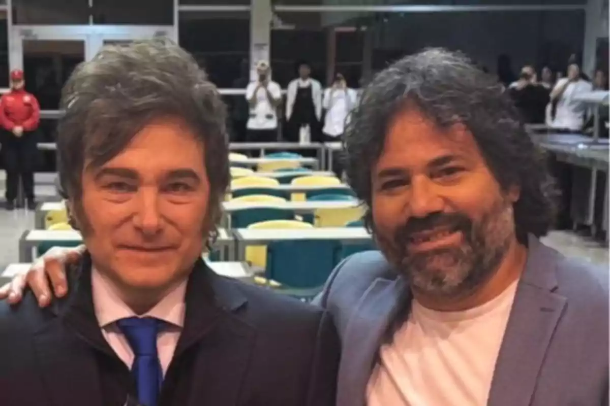 Dos hombres posan juntos en un comedor con mesas y sillas de colores al fondo, ambos sonríen y visten saco