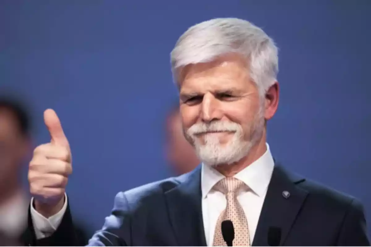 Hombre de cabello y barba canosos usando traje y corbata sonríe y levanta el pulgar en señal de aprobación frente a un fondo azul