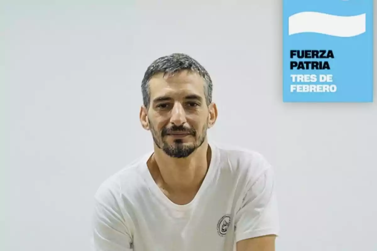 Short-haired, bearded man wearing a white t-jersey sitting in front of a white wall with a blue sign that says Fuerza Patria Tres de Febrero