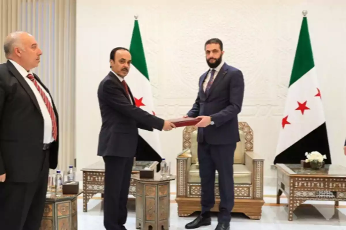 Tres hombres con trajes formales participan en una ceremonia oficial en una sala elegante con dos banderas de fondo y muebles decorativos
