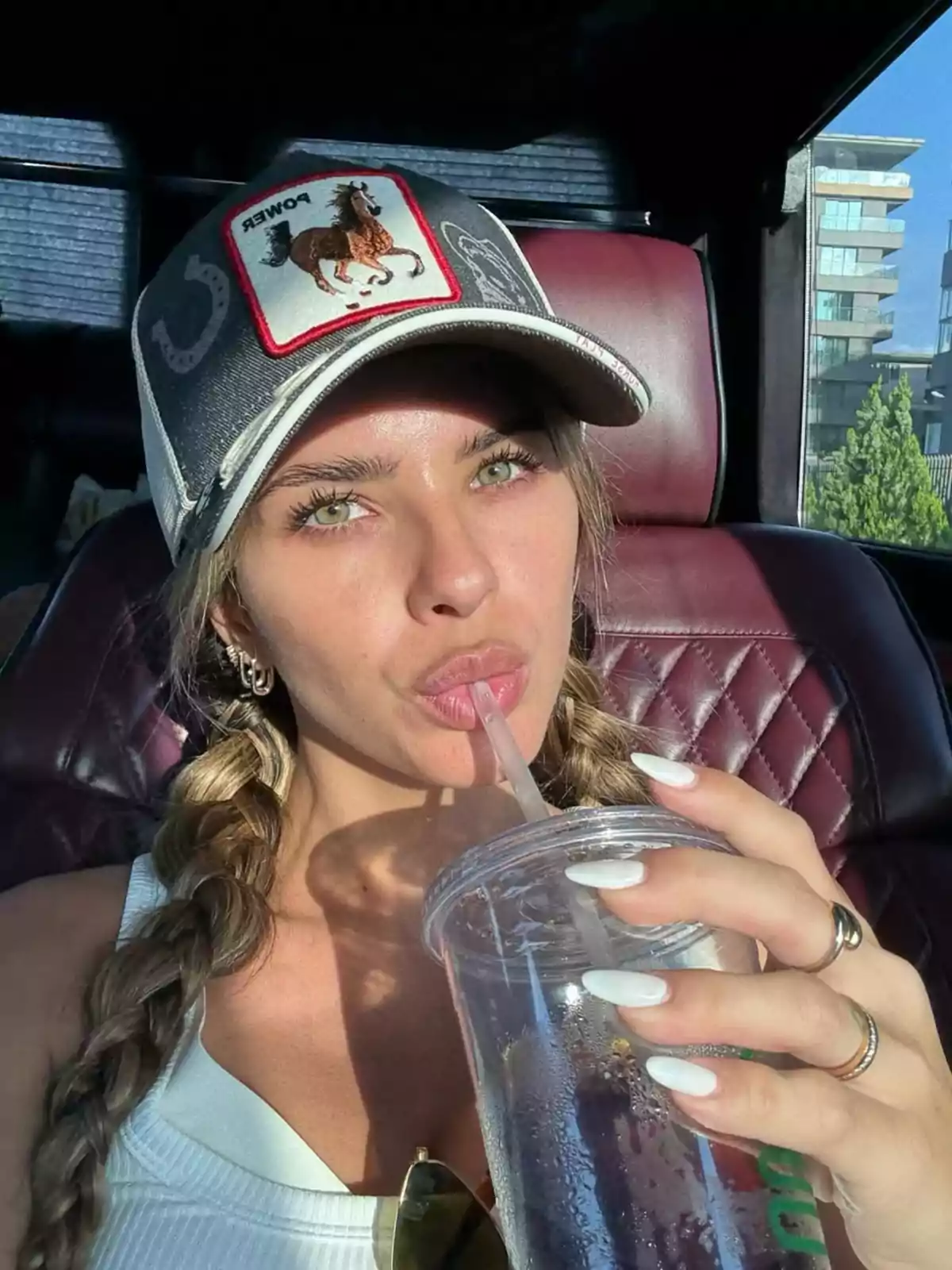 Woman with braids and a cap with an embroidered horse, drinking from a clear glass inside a car Woman with braids and a cap with an embroidered horse, drinking from a clear glass inside a car