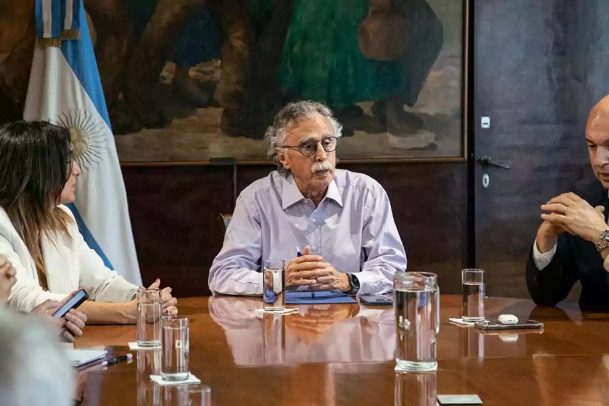 Un hombre mayor con gafas y bigote está sentado en una mesa de reuniones junto a otras personas, con vasos de agua y una bandera de Argentina al fondo