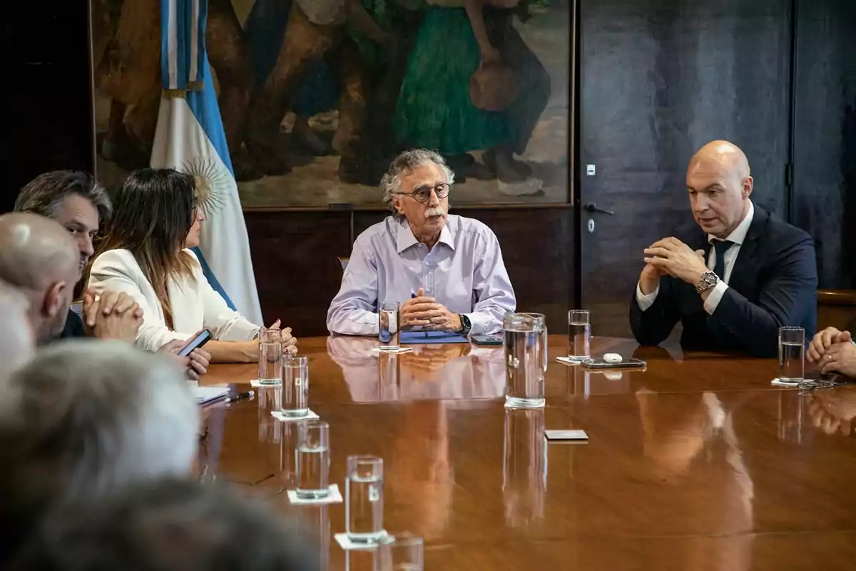 Varias personas sentadas alrededor de una mesa de madera en una reunión formal con vasos de agua y una bandera de Argentina al fondo Varias personas sentadas alrededor de una mesa de madera en una reunión formal con vasos de agua y una bandera de Argentina al fondo