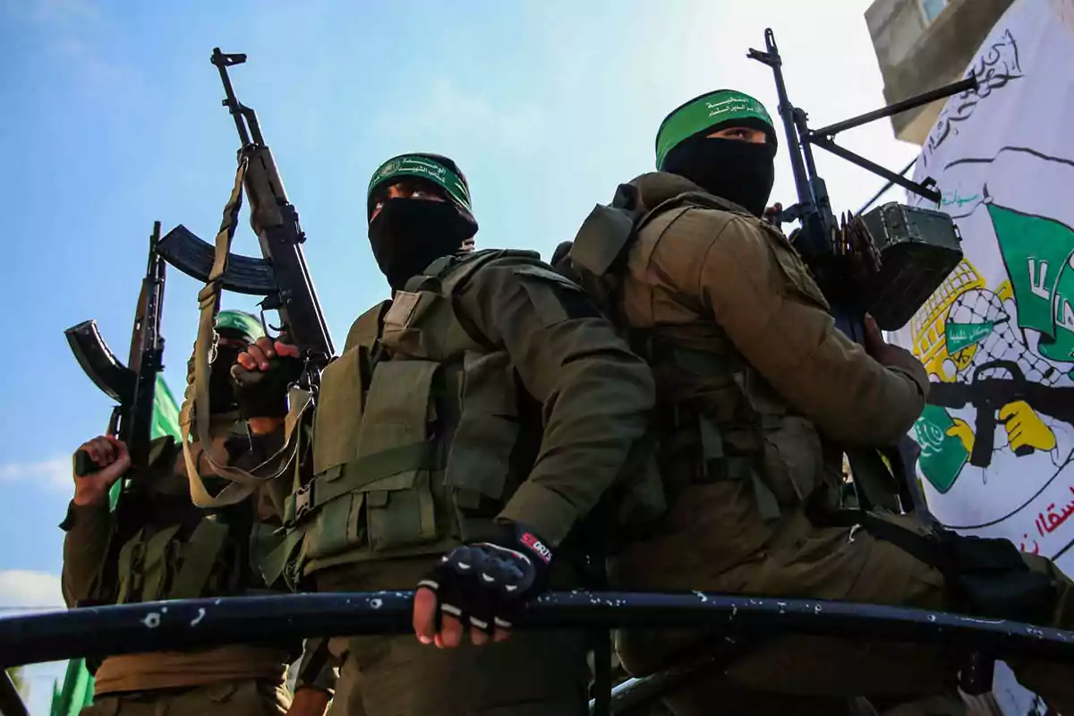 Tres personas con uniformes militares y pasamontañas portan armas largas y están de pie junto a una bandera con símbolos y letras árabes. Tres personas con uniformes militares y pasamontañas portan armas largas y están de pie junto a una bandera con símbolos y letras árabes.