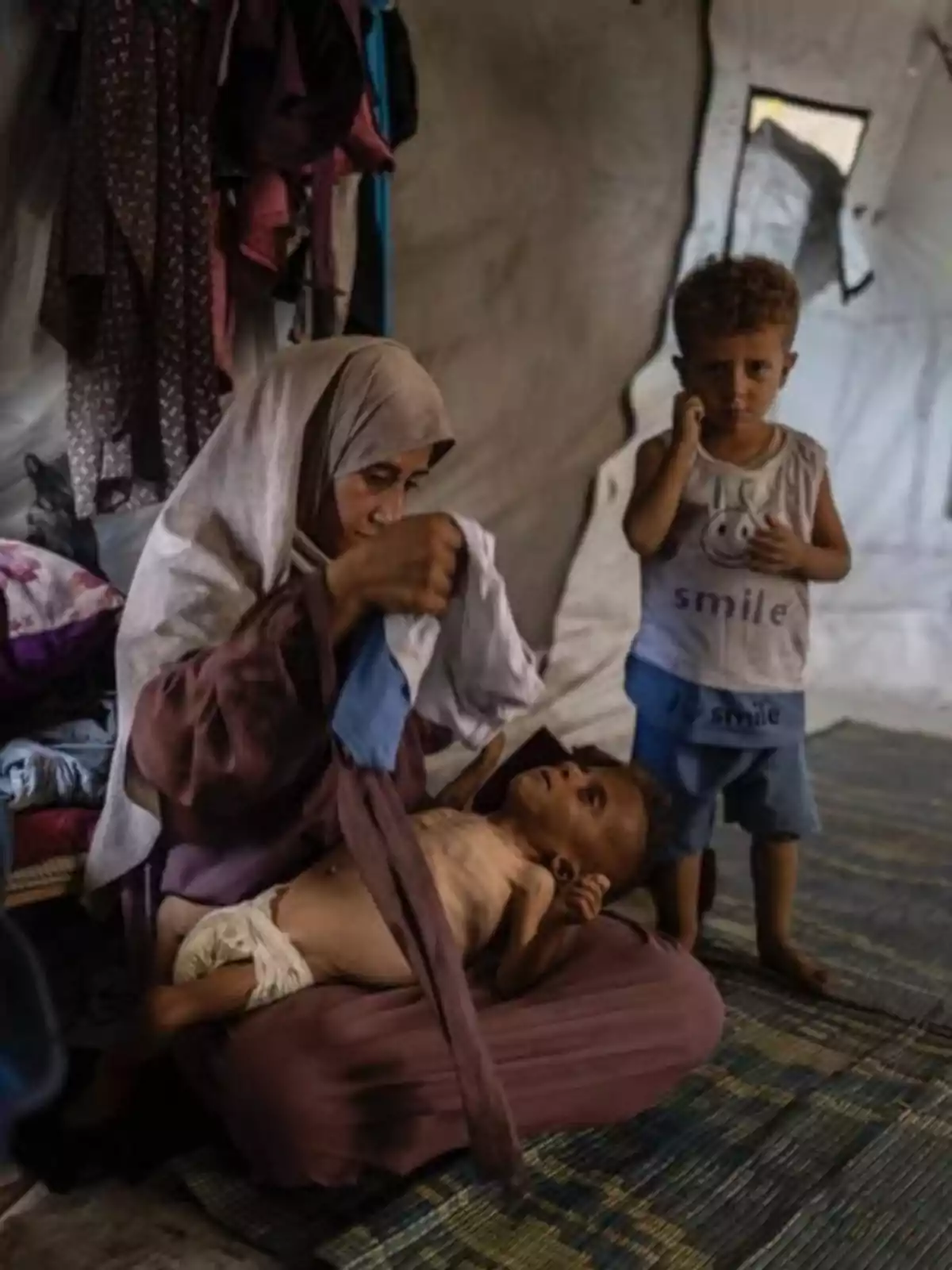 A veiled woman tends to a small child lying across her lap inside a tent while another child stands by her side watching. A veiled woman tends to a small child lying across her lap inside a tent while another child stands by her side watching.