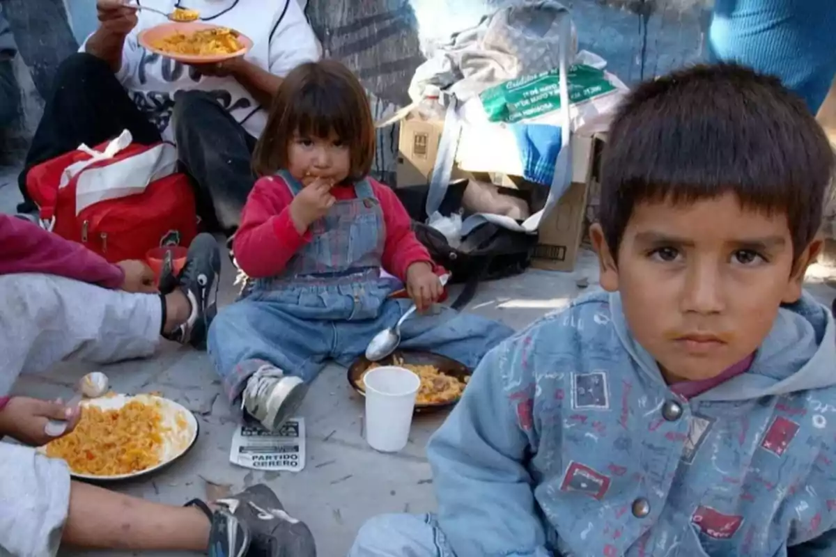 Niños sentados en el suelo comiendo comida de platos y vasos desechables en un entorno humilde Niños sentados en el suelo comiendo comida de platos y vasos desechables en un entorno humilde