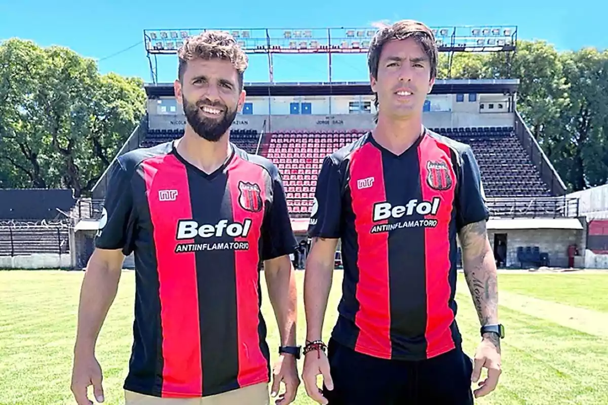 Two men wearing red and black striped soccer jerseys pose on a field with bleachers and trees in the background. Two men wearing red and black striped soccer jerseys pose on a field with bleachers and trees in the background.