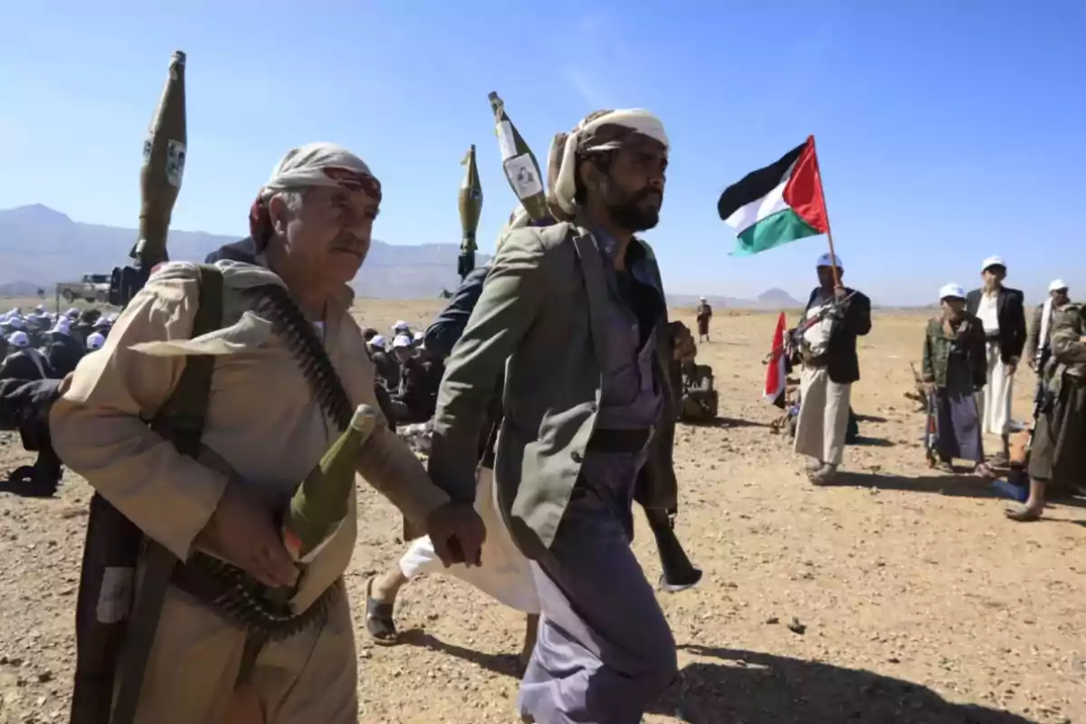 Hombres armados con vestimenta tradicional yemení caminan en un terreno desértico mientras al fondo se observa un grupo de personas y una bandera palestina ondeando