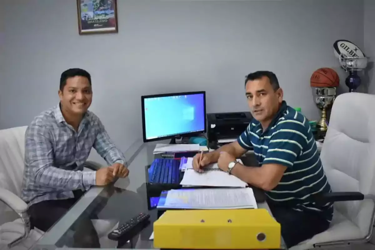 Two men sitting in an office across from a desk with documents, a computer, and trophies in the background. Two men sitting in an office across from a desk with documents, a computer, and trophies in the background.