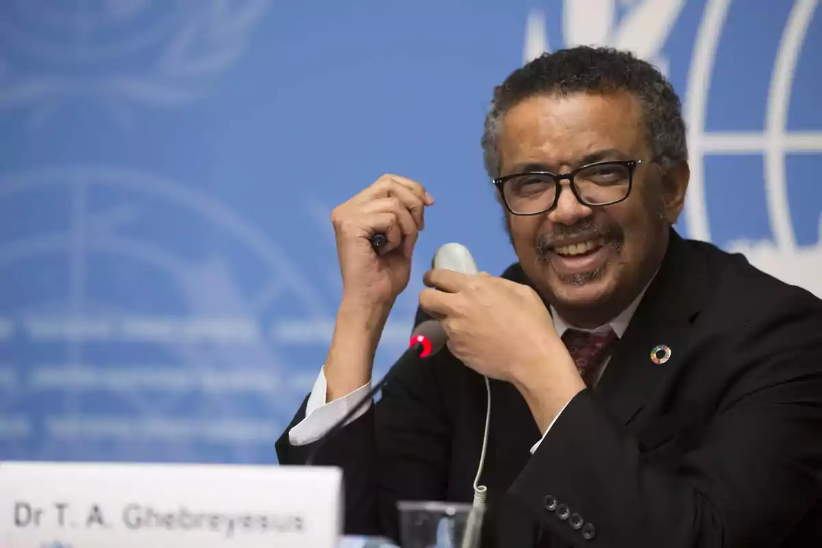 Hombre con gafas y traje oscuro sonriendo mientras sostiene un micrófono en una conferencia con fondo azul y logotipo institucional. Hombre con gafas y traje oscuro sonriendo mientras sostiene un micrófono en una conferencia con fondo azul y logotipo institucional.