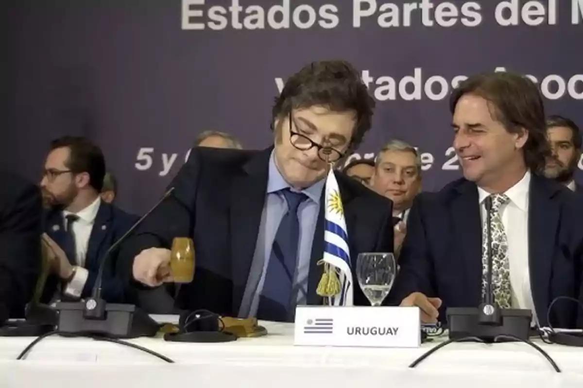 Un hombre con gafas sostiene un mazo junto a una bandera de Uruguay en una mesa de conferencia, mientras otro hombre a su lado sonríe, rodeados de otras personas en un evento formal. Un hombre con gafas sostiene un mazo junto a una bandera de Uruguay en una mesa de conferencia, mientras otro hombre a su lado sonríe, rodeados de otras personas en un evento formal.