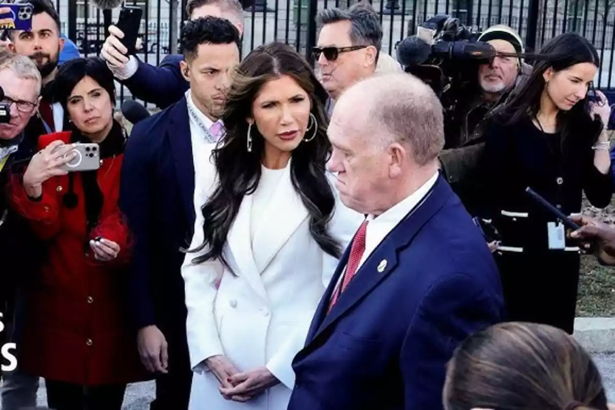 Una mujer vestida de blanco y un hombre de traje azul rodeados de periodistas y cámaras en un evento al aire libre Una mujer vestida de blanco y un hombre de traje azul rodeados de periodistas y cámaras en un evento al aire libre