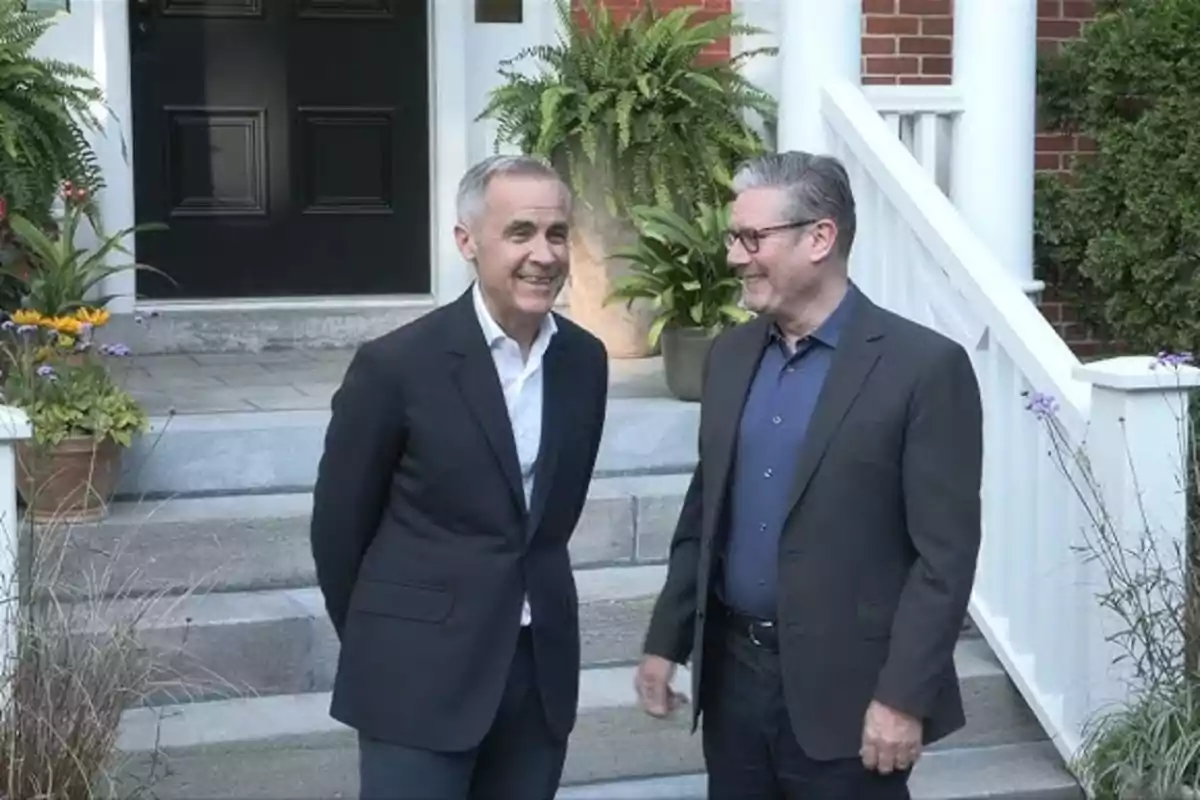 Dos hombres de traje conversan y sonríen frente a la entrada de una casa con plantas y escalones.