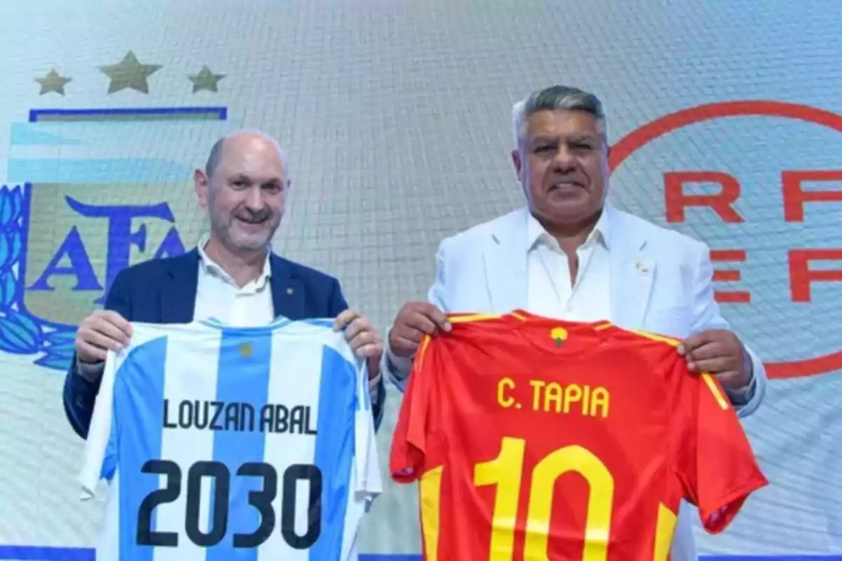 Dos hombres posan sonrientes sosteniendo camisetas de fútbol personalizadas, una de Argentina y otra de España, frente a los logotipos de la AFA y la RFEF. Dos hombres posan sonrientes sosteniendo camisetas de fútbol personalizadas, una de Argentina y otra de España, frente a los logotipos de la AFA y la RFEF.