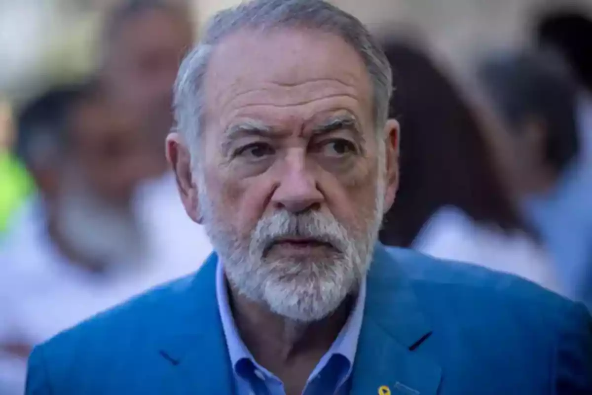 Older man with gray hair and beard wearing a blue jacket with a serious expression in an outdoor setting with blurred people in the background