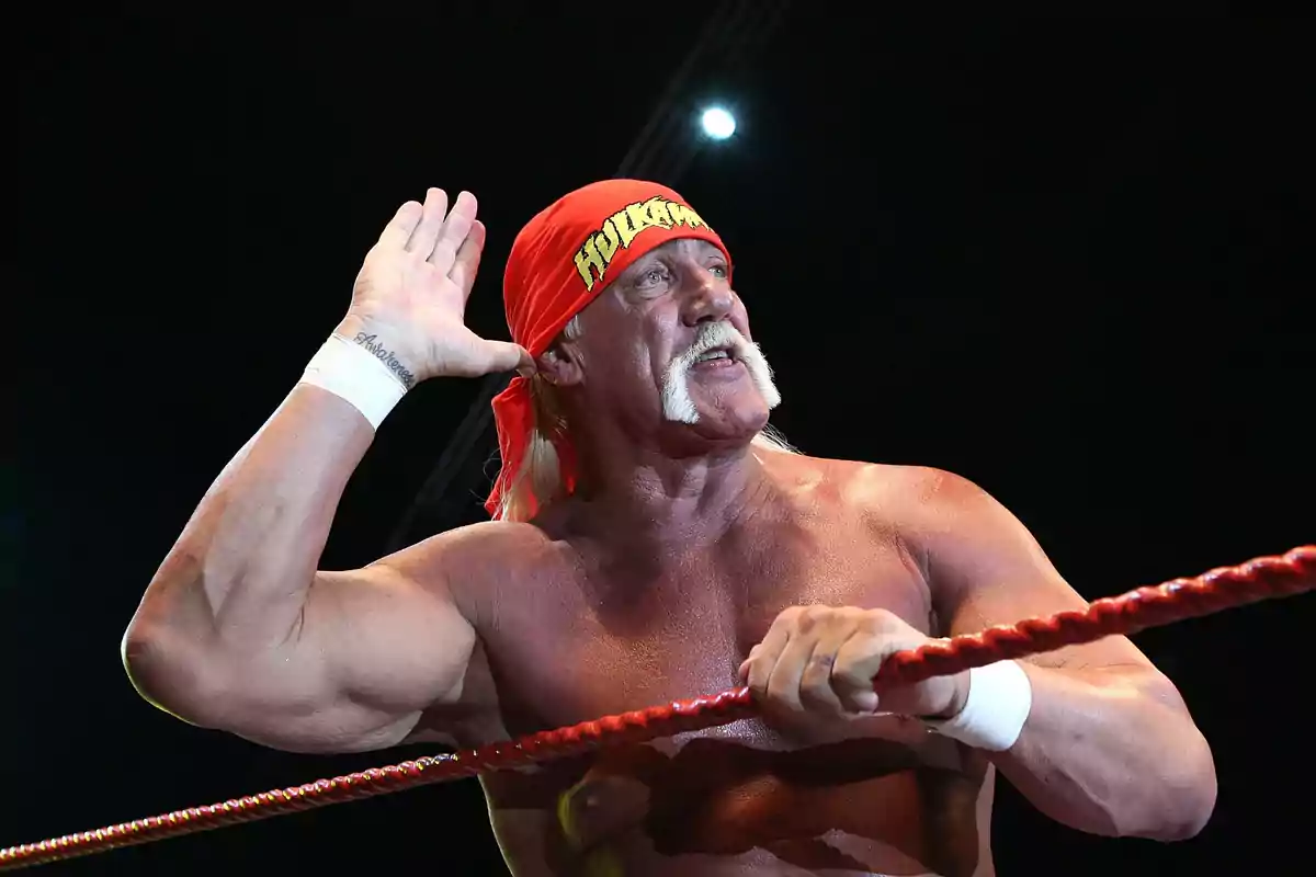 Muscular wrestler with a red bandana on his head and a white mustache posing in a wrestling ring while holding his hand to his ear