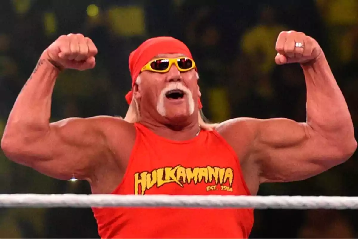 Hombre musculoso con camiseta roja y gafas de sol amarillas levantando ambos brazos en señal de fuerza dentro de un ring de lucha libre