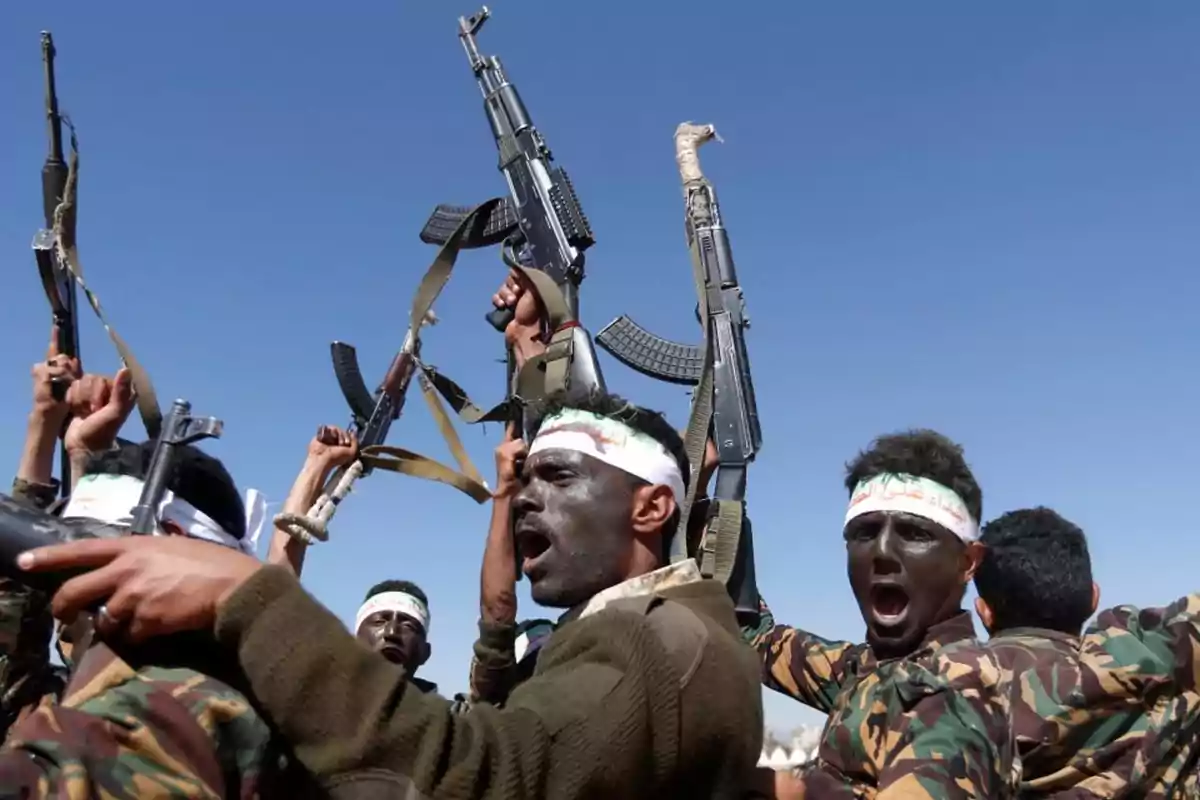Grupo de hombres con la cara pintada y cintas en la cabeza levantando rifles al aire bajo un cielo despejado Grupo de hombres con la cara pintada y cintas en la cabeza levantando rifles al aire bajo un cielo despejado