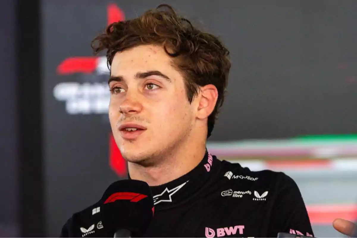 Motorsport driver in a black sponsor-branded uniform speaking into a microphone at a press conference Motorsport driver in a black sponsor-branded uniform speaking into a microphone at a press conference