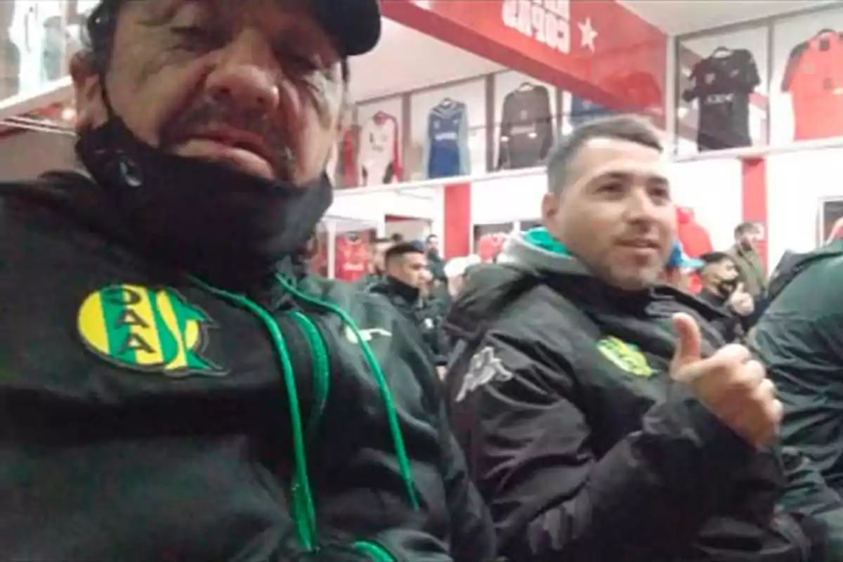 Dos hombres con ropa deportiva de un club de fútbol posan en un salón con camisetas en exhibición al fondo, uno de ellos muestra el pulgar hacia arriba. Dos hombres con ropa deportiva de un club de fútbol posan en un salón con camisetas en exhibición al fondo, uno de ellos muestra el pulgar hacia arriba.