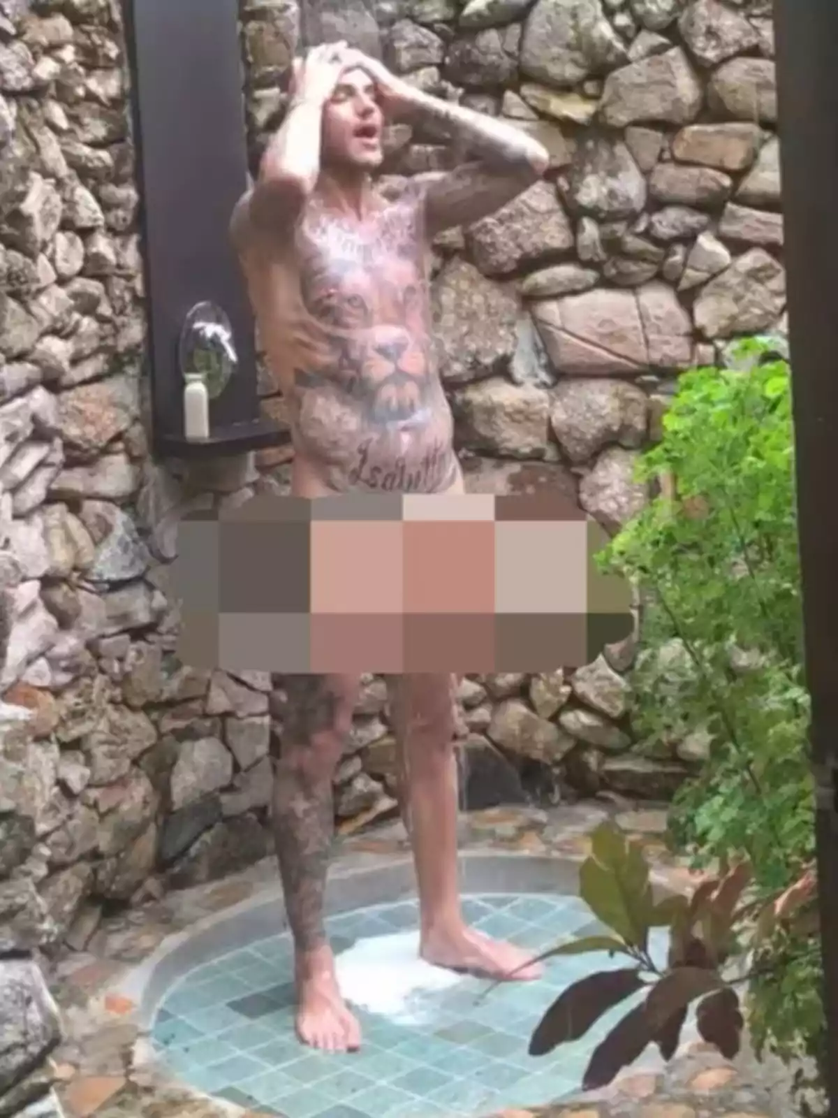 Hombre tatuado tomando una ducha al aire libre en un espacio rodeado de paredes de piedra con vegetación cercana y una zona pixelada cubriendo la parte inferior de su cuerpo Hombre tatuado tomando una ducha al aire libre en un espacio rodeado de paredes de piedra con vegetación cercana y una zona pixelada cubriendo la parte inferior de su cuerpo