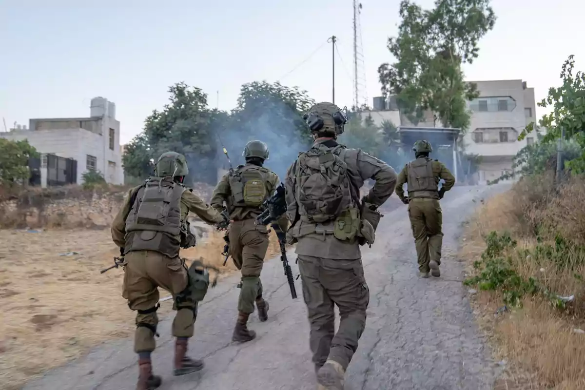 Four armed soldiers in military uniforms advance along a dirt road in an urban area surrounded by vegetation and buildings. Four armed soldiers in military uniforms advance along a dirt road in an urban area surrounded by vegetation and buildings.