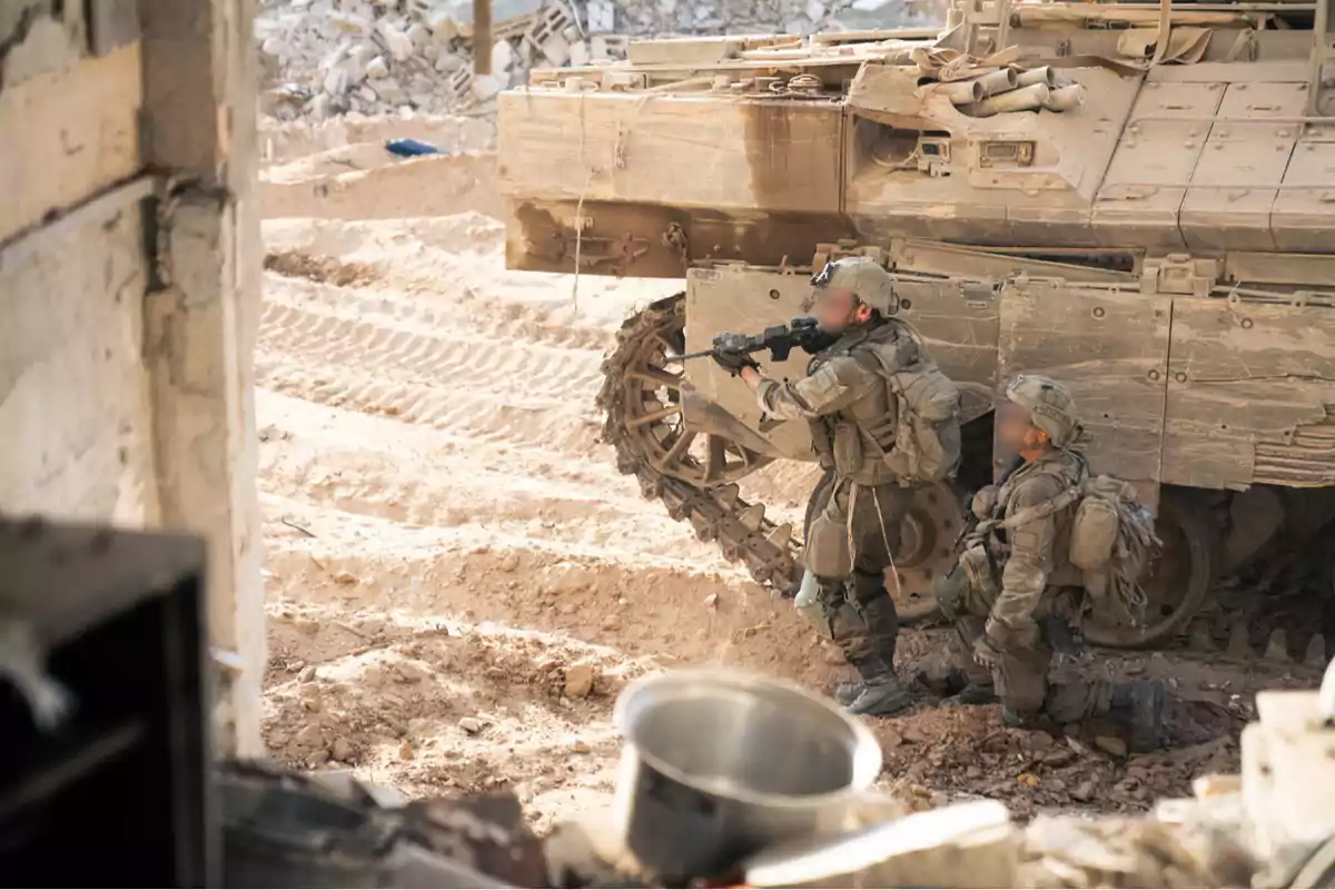 Two armed soldiers in military uniforms and tactical gear take cover next to an armored vehicle in an area of rubble and dirt.