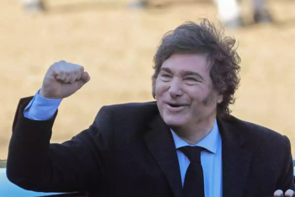 Hombre de cabello despeinado con traje oscuro levantando el puño y sonriendo en un ambiente al aire libre