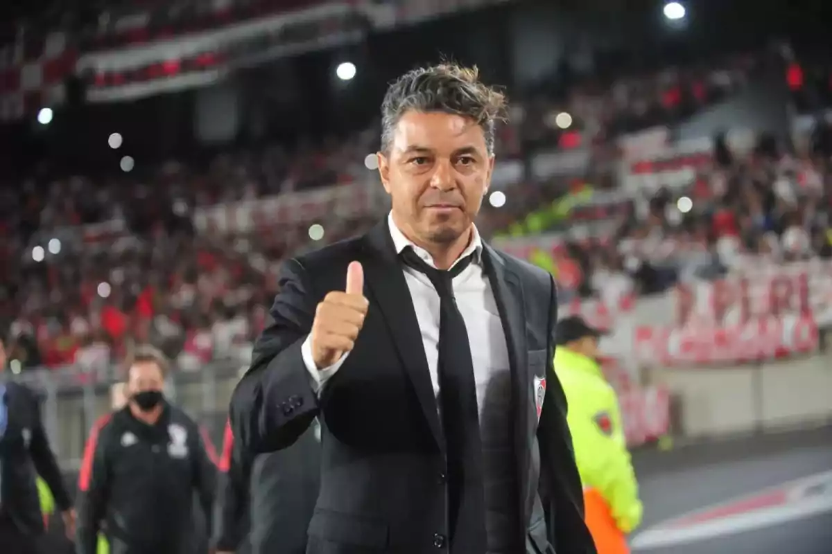 Hombre con traje oscuro y corbata mostrando el pulgar hacia arriba en un estadio lleno de gente