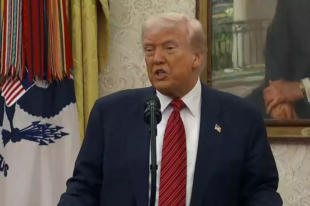 Blond-haired man in a blue suit and red tie speaking in front of a microphone in an official room decorated with flags and a painting in the background Blond-haired man in a blue suit and red tie speaking in front of a microphone in an official room decorated with flags and a painting in the background