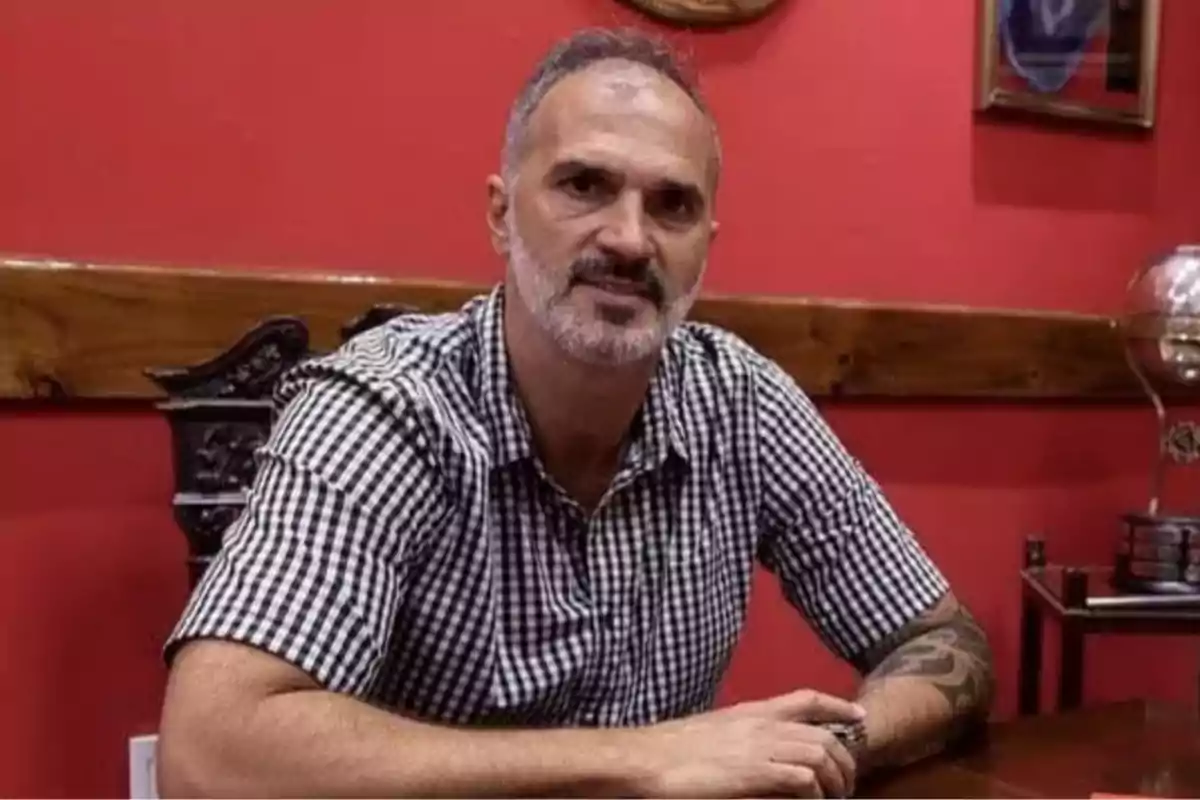 Hombre de cabello corto y barba sentado en una mesa con camisa de cuadros en una habitación de paredes rojas