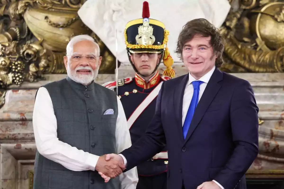 Two men in suits shake hands in front of a guard in ceremonial uniform and a decorative golden background.