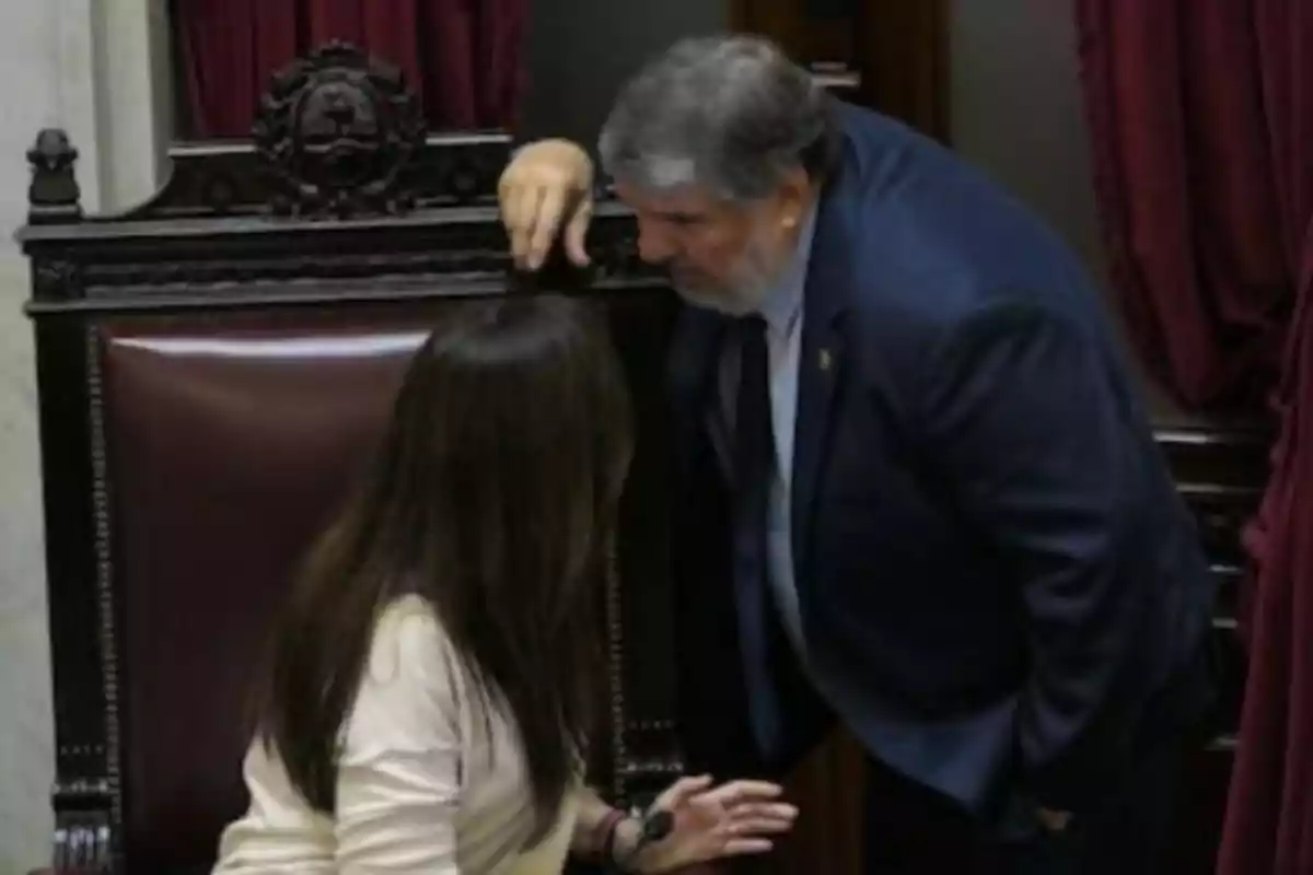 A man in a suit leans down to speak quietly to a woman seated in a large chair inside a parliamentary chamber.