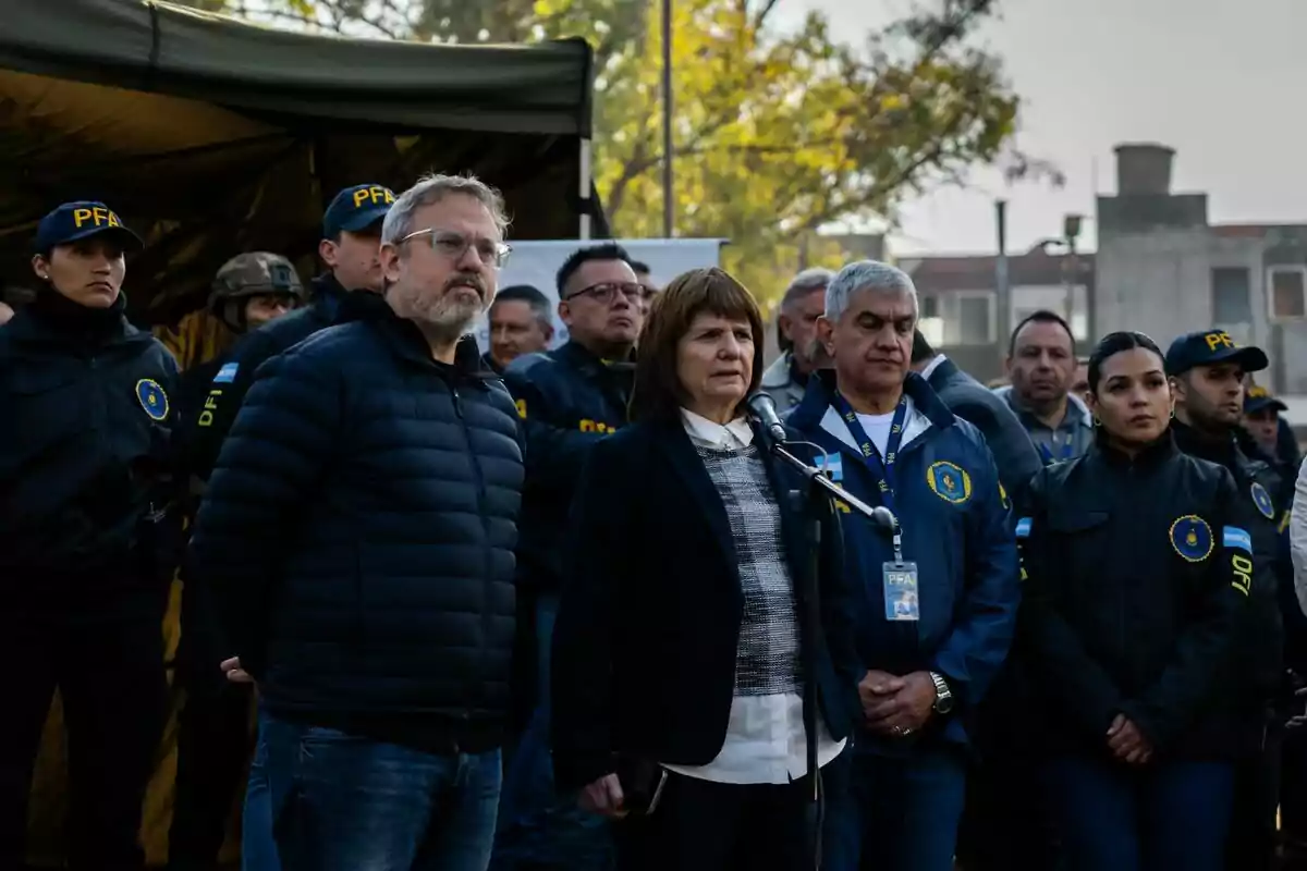 Un grupo de personas con uniformes policiales y credenciales oficiales se encuentra reunido al aire libre mientras una mujer habla frente a un micrófono