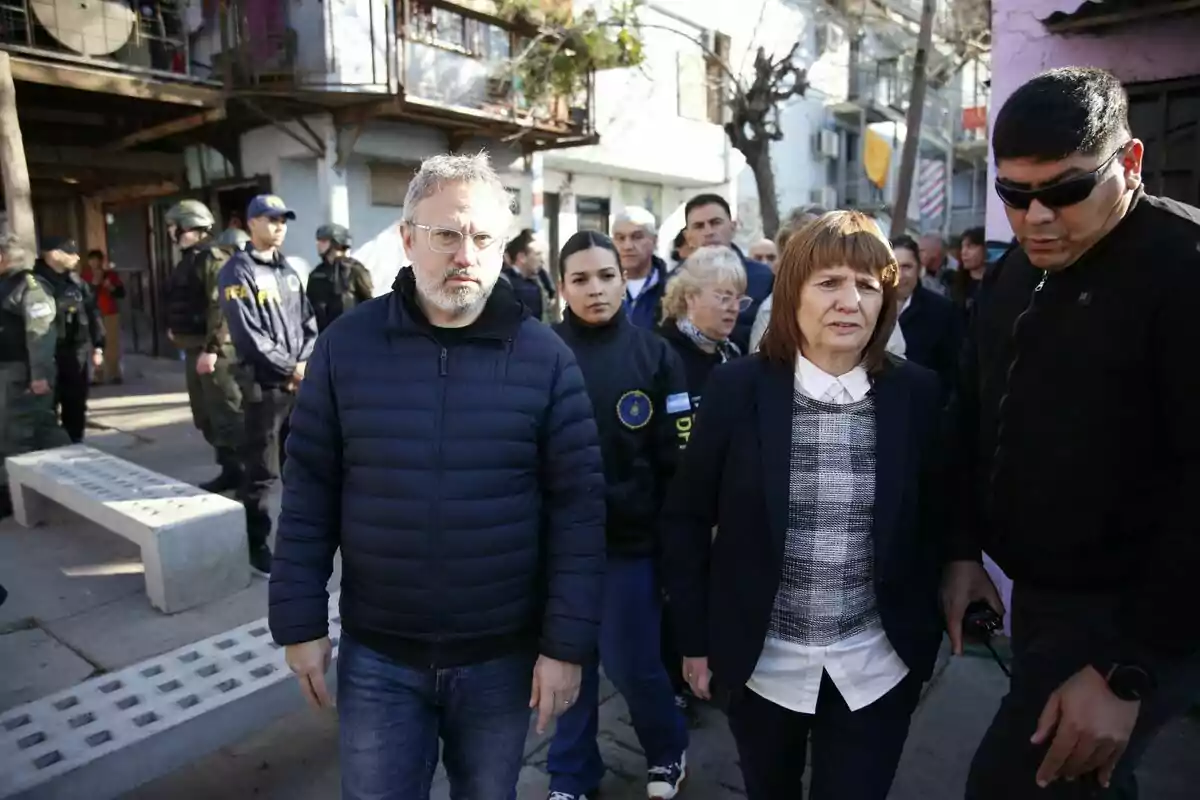 Un grupo de personas camina por una calle en un barrio, acompañados por fuerzas de seguridad y rodeados de edificios y vecinos. Un grupo de personas camina por una calle en un barrio, acompañados por fuerzas de seguridad y rodeados de edificios y vecinos.