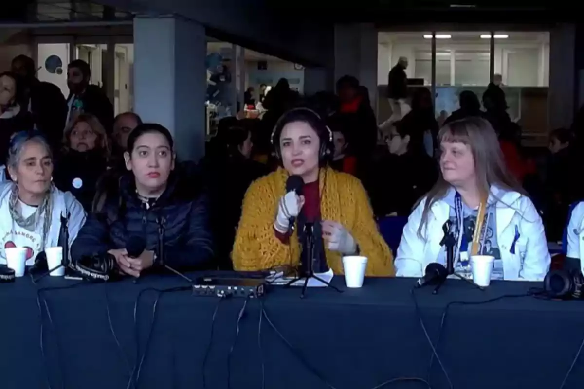 Cuatro mujeres sentadas en una mesa con micrófonos y auriculares, participando en una conferencia o transmisión en un espacio cerrado con varias personas al fondo.