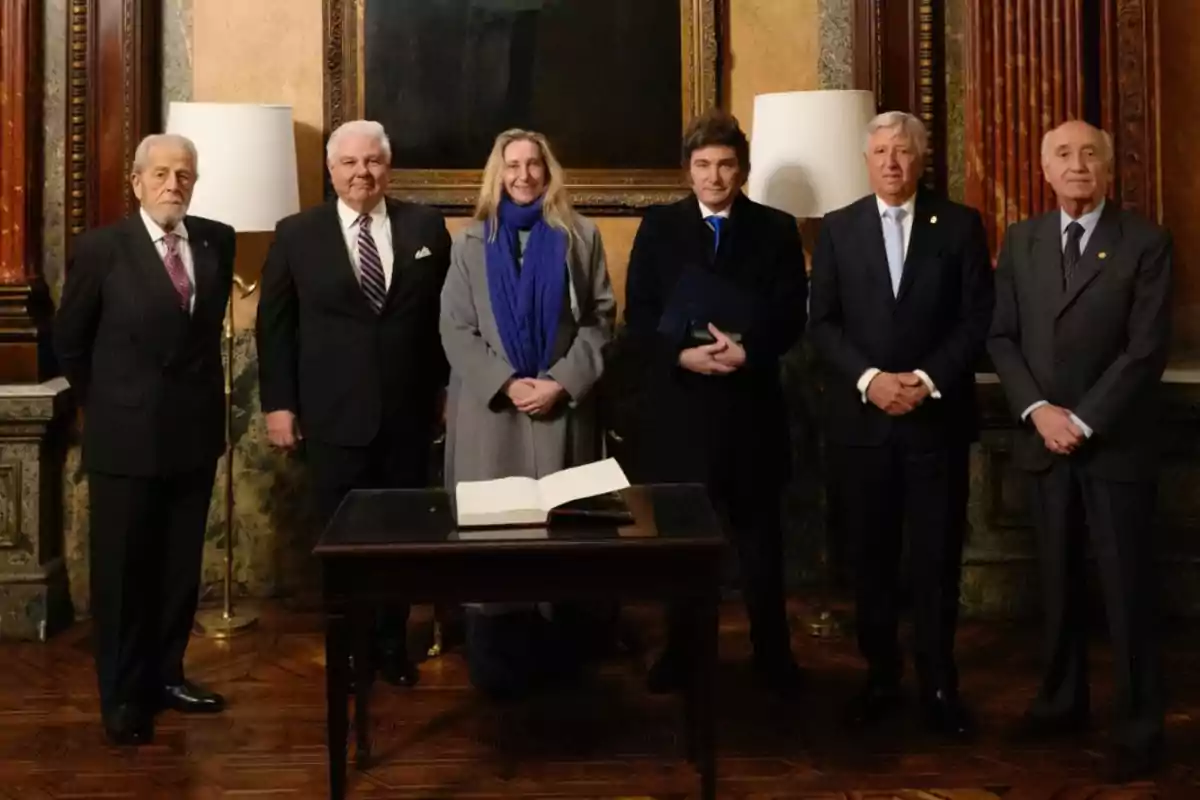 Six people stand posing in an elegant room with decorated walls and a table with an open book in front. Six people stand posing in an elegant room with decorated walls and a table with an open book in front.