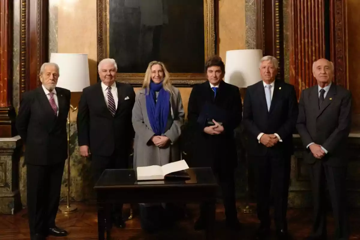 Six people stand posing in an elegant hall with marble and wood walls, behind a table with an open book. Six people stand posing in an elegant hall with marble and wood walls, behind a table with an open book.