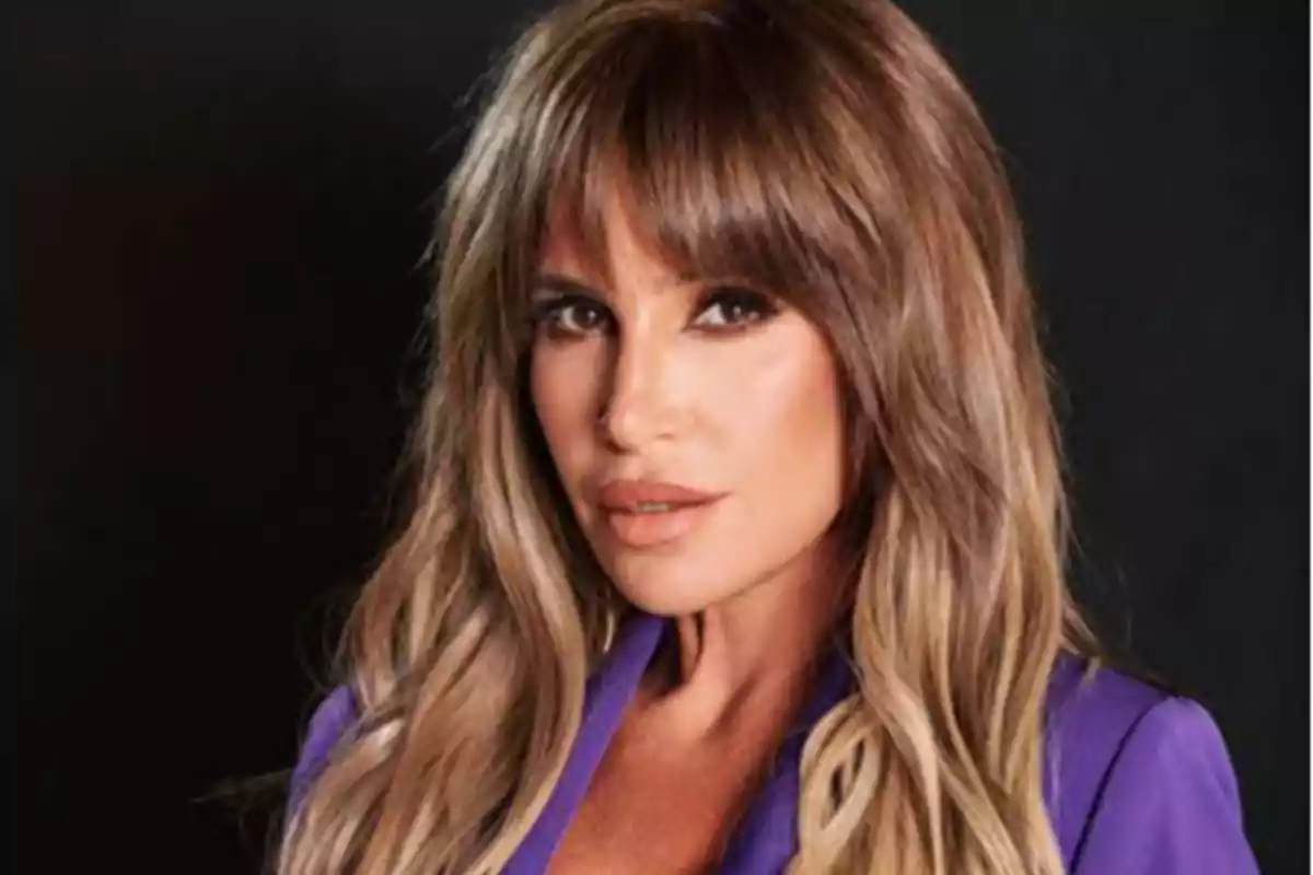 Mujer de cabello largo y rubio con flequillo usando un saco morado y fondo oscuro