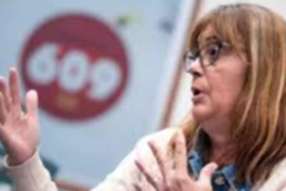 Mujer de cabello castaño y gafas gesticulando con las manos frente a un fondo borroso donde se distingue un círculo rojo con el número 609 Mujer de cabello castaño y gafas gesticulando con las manos frente a un fondo borroso donde se distingue un círculo rojo con el número 609