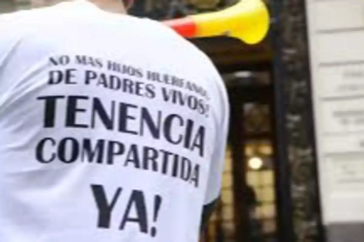 Persona de espaldas con camiseta blanca que lleva un mensaje a favor de la tenencia compartida y sostiene una corneta amarilla Persona de espaldas con camiseta blanca que lleva un mensaje a favor de la tenencia compartida y sostiene una corneta amarilla