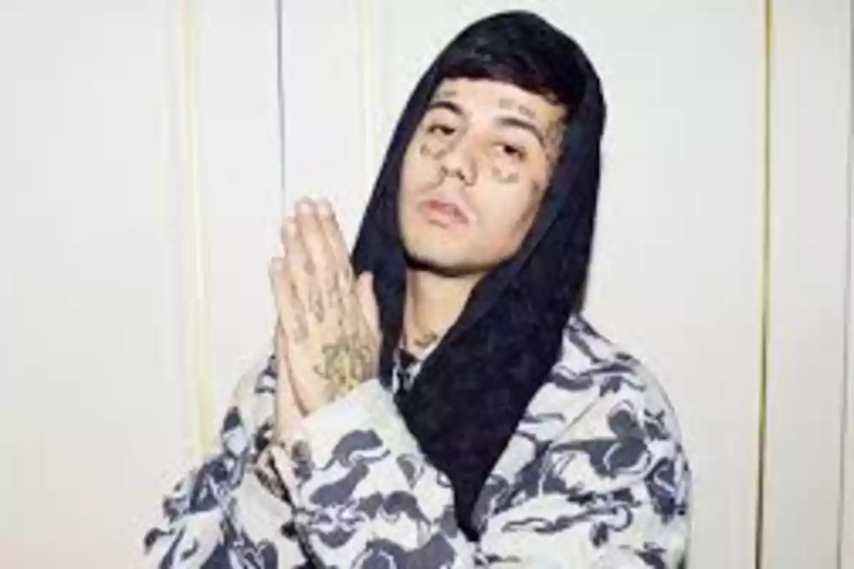 Young man wearing a camouflage print hoodie with a black hood, tattoos on his face, and hands clasped in prayer in front of a light-colored wall Young man wearing a camouflage print hoodie with a black hood, tattoos on his face, and hands clasped in prayer in front of a light-colored wall