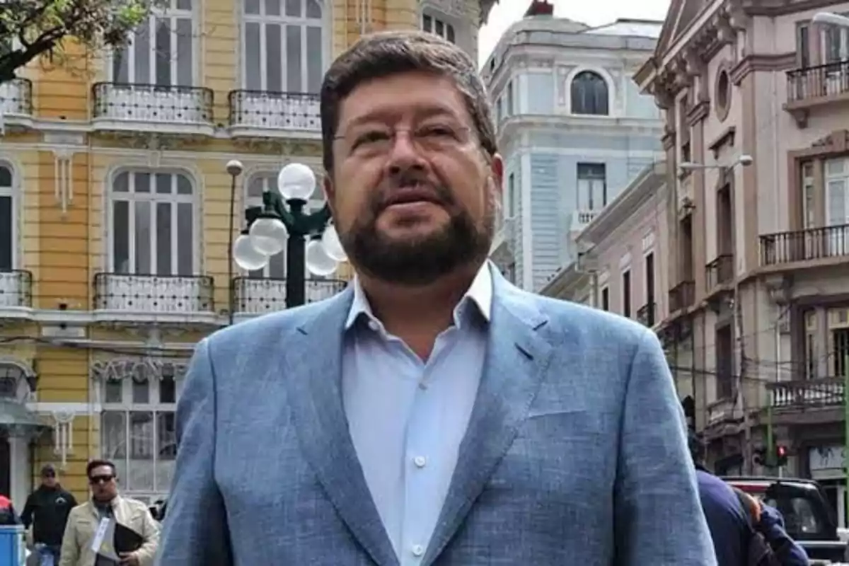 Hombre con barba y gafas usando saco azul claro de pie en una calle con edificios antiguos de fondo