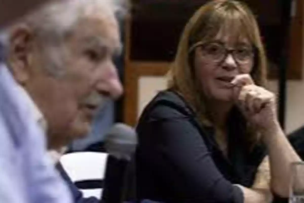Una mujer con gafas observa atentamente a un hombre mayor que sostiene un micrófono durante una conversación en un ambiente interior Una mujer con gafas observa atentamente a un hombre mayor que sostiene un micrófono durante una conversación en un ambiente interior