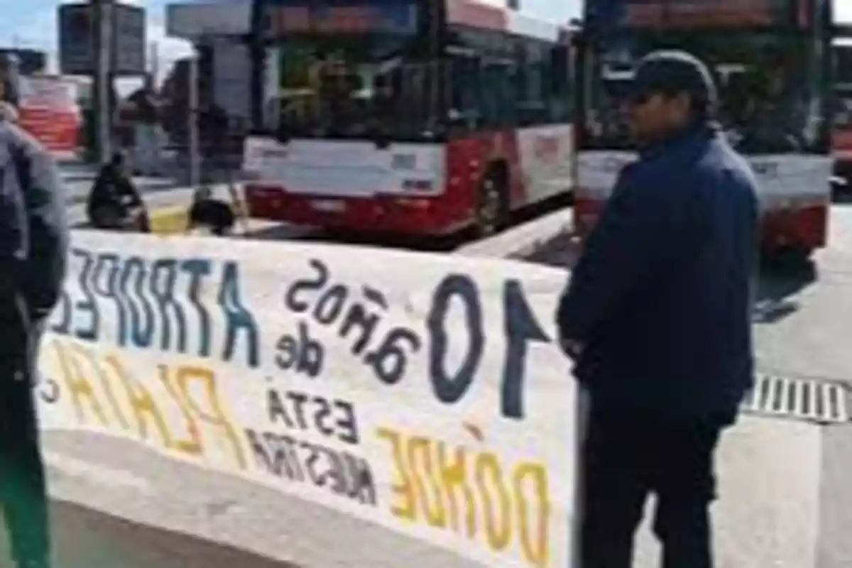 Personas sostienen una manta de protesta frente a varios autobuses rojos en una calle urbana Personas sostienen una manta de protesta frente a varios autobuses rojos en una calle urbana