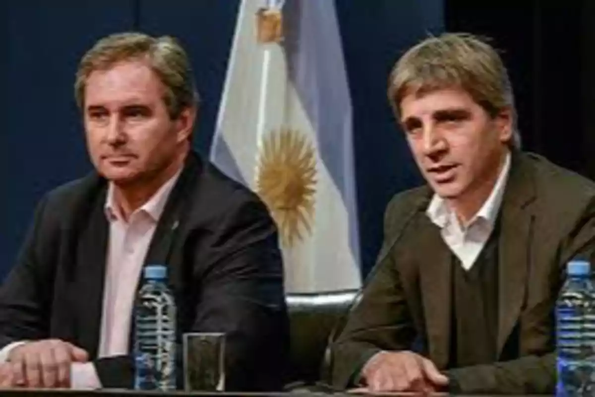 Dos hombres con traje sentados en una mesa durante una conferencia de prensa con una bandera de Argentina de fondo y botellas de agua sobre la mesa